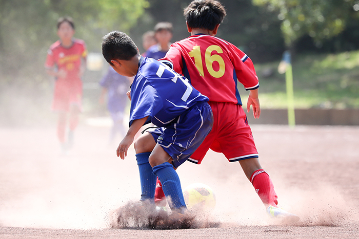 土のグラウンドでサッカーをする小学生がボールを奪い合う場面